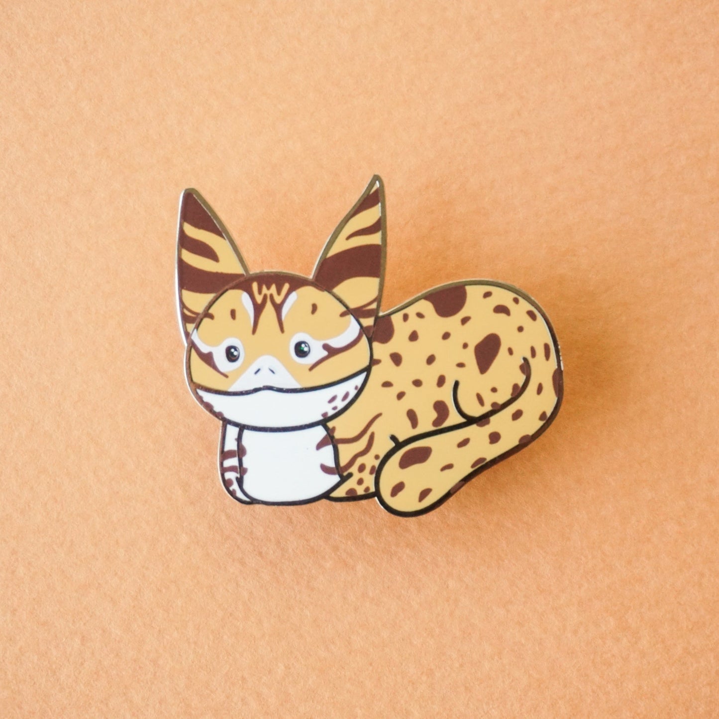 A loafing lothcat enamel pin in tan and brown and white colors.