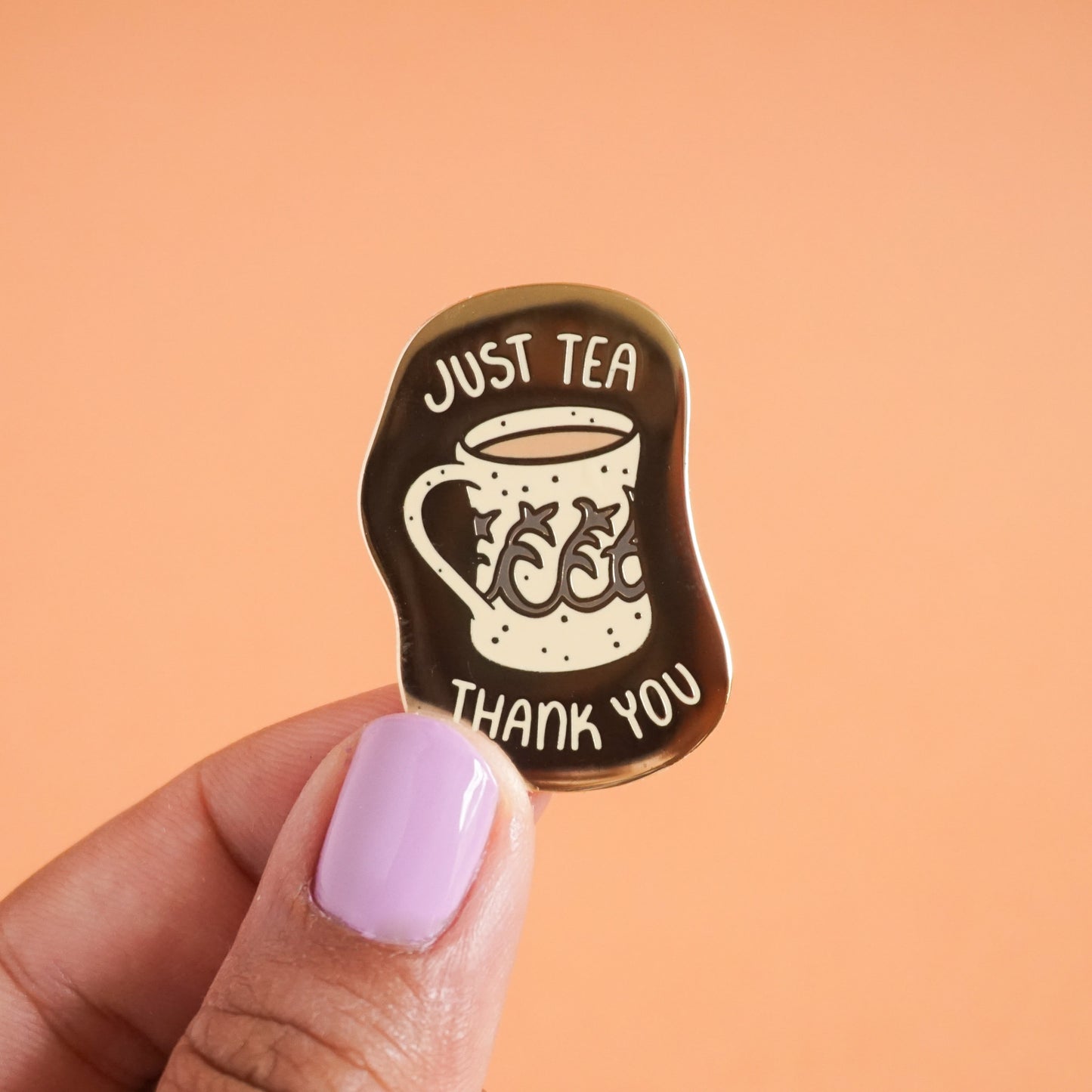 A small gold enamel pin with the words "just tea thank you" on it around a vintage style mug with tea in it.