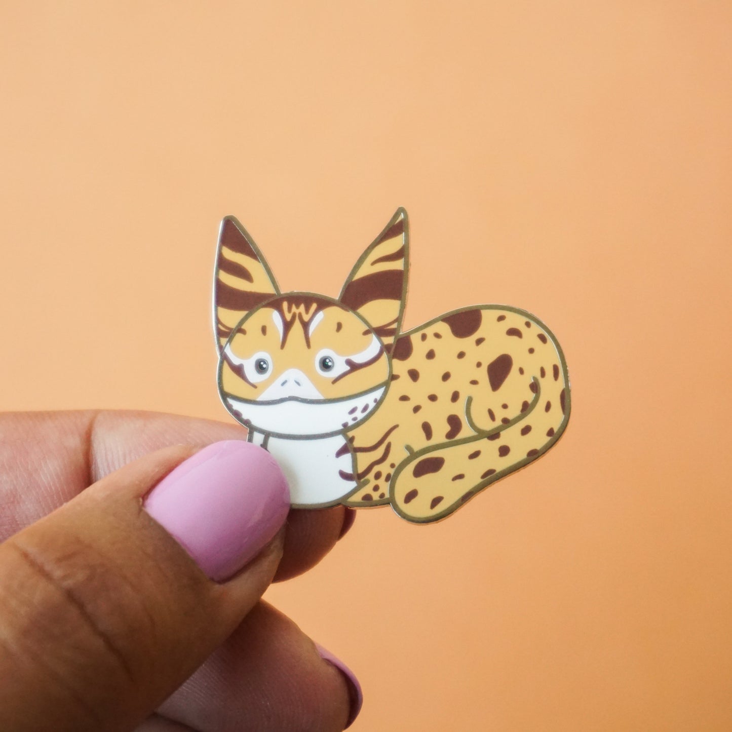 A loafing lothcat enamel pin in tan and brown and white colors.