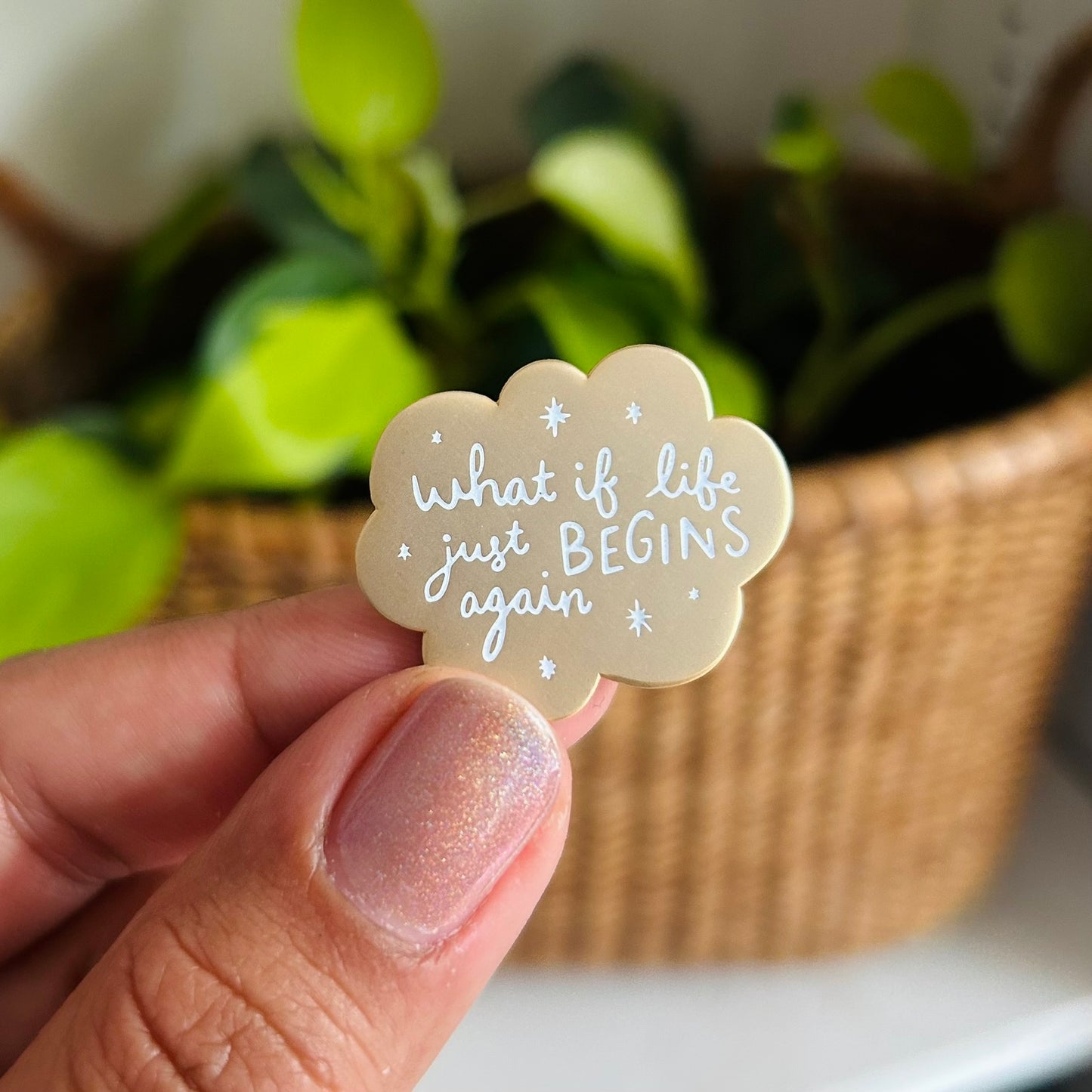 A hand holds up a small matte gold enamel pin with scalloped edges and white stars around the words "what if life just begins again?"
