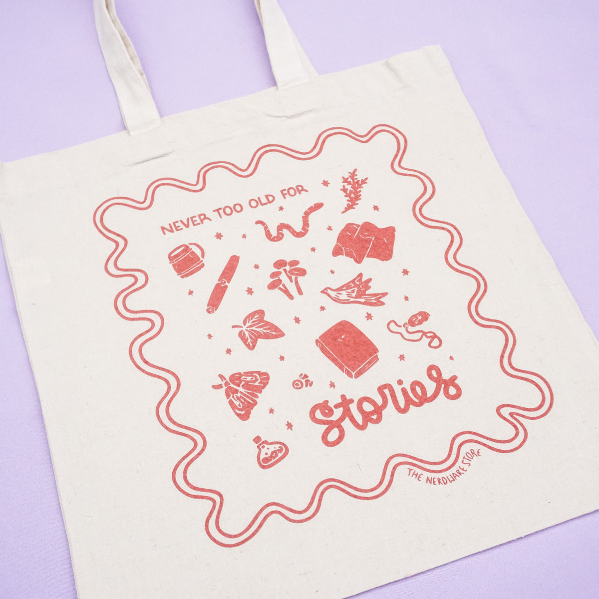 tote bag with designs in maroon that say never too old for stories with fantastical illustrations of a book, locket, moth, leaf, and more