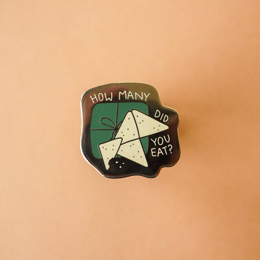 A small gold enamel pin that says "how many did you eat?" around an open packet of lembas bread from the Lord of the Rings.