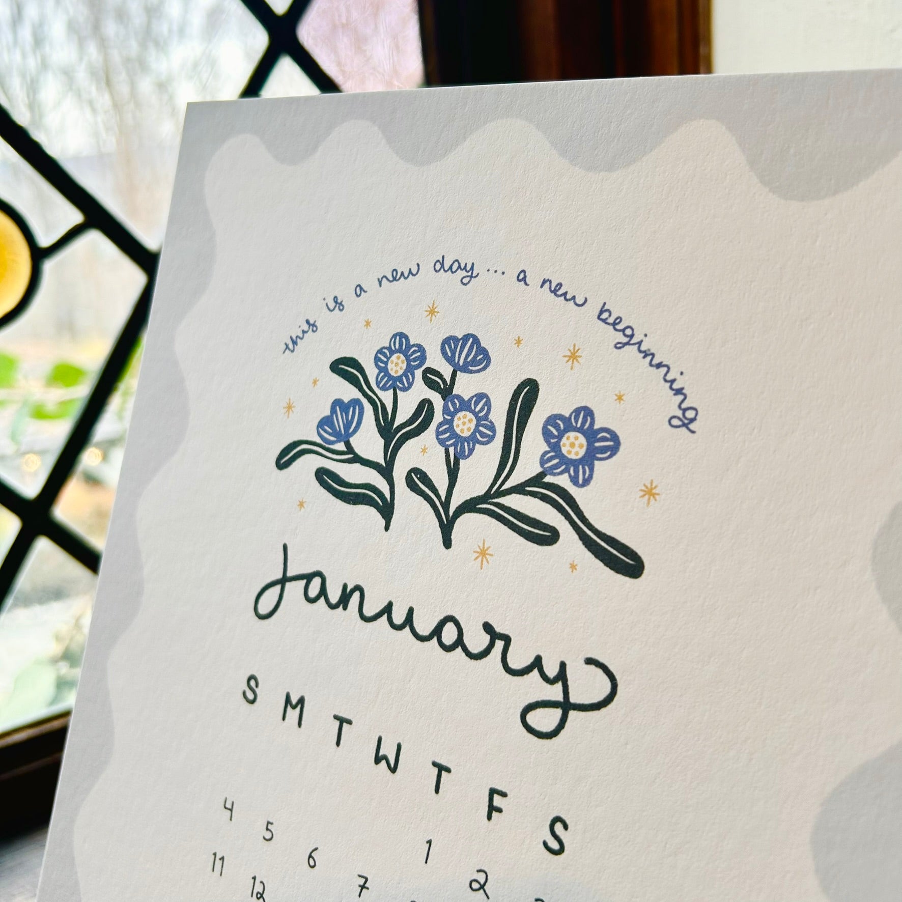 calendar page for july with forget-me-nots and the words "this is a new day...a new beginning - the calendar sits in front of a diamond pane window.