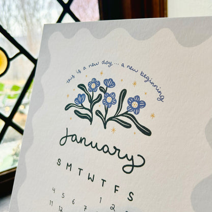 calendar page for july with forget-me-nots and the words "this is a new day...a new beginning - the calendar sits in front of a diamond pane window.