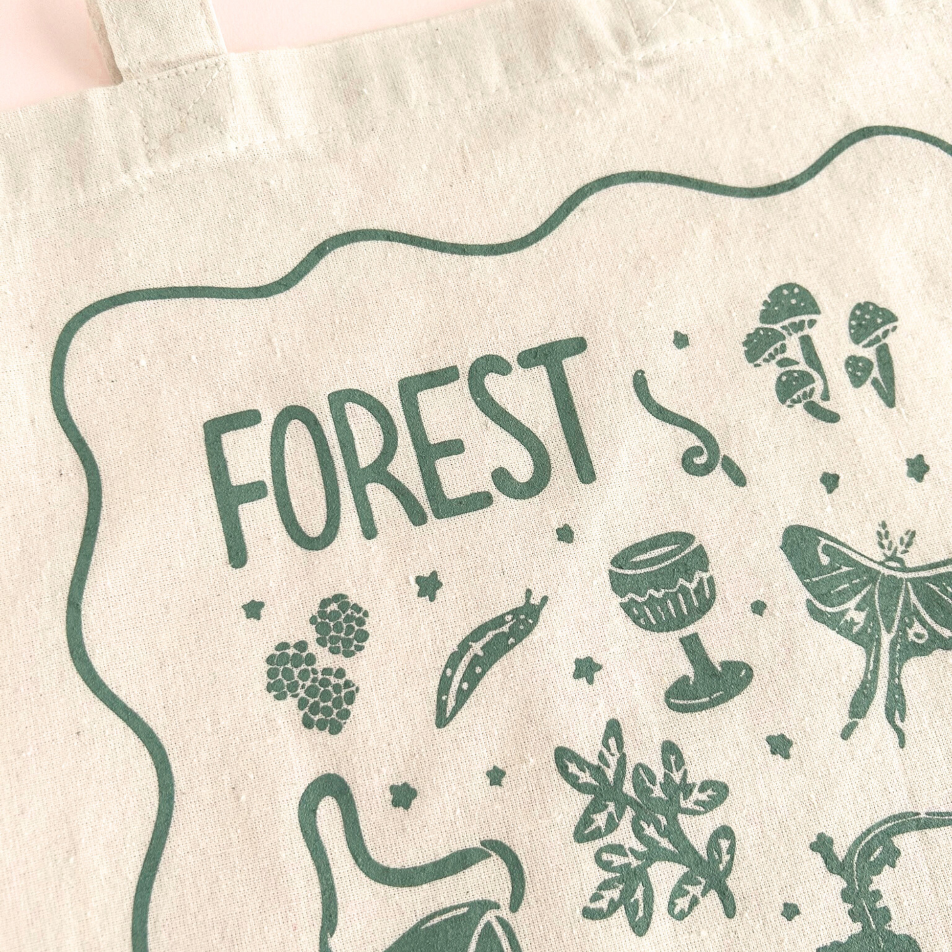 tote bag with designs in dark green that say forest goblin with fantastical illustrations of a slug, a goblet, a satchel, foliage, a lantern, mushrooms, and a luna moth