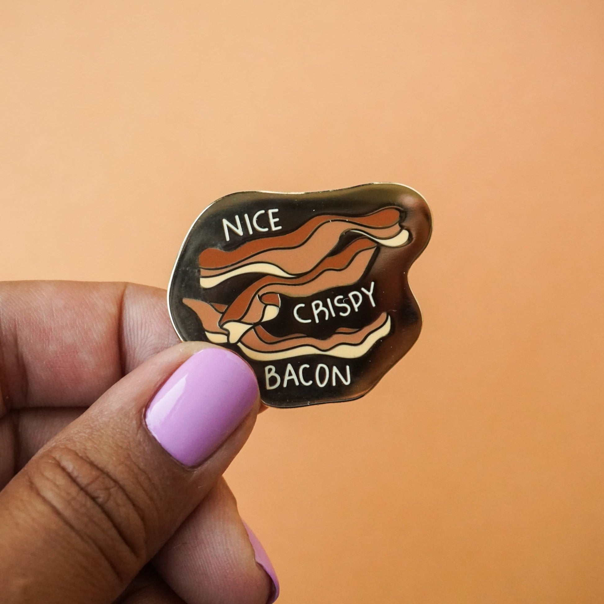 A small gold enamel pin with bacon on it and the words "nice, crispy, bacon."