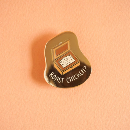 A gold enamel pin with a salt box open on the pin and the words "roast chicken?" around the salt box.