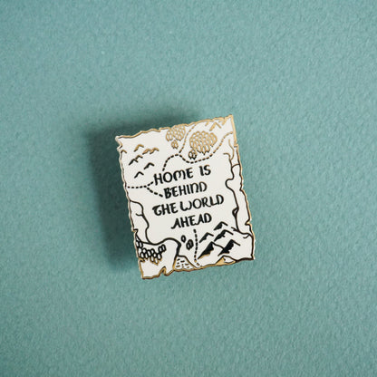 A small gold enamel pin of a map in the style of the lord of the rings with the words "Home is behind, the world ahead" on it.