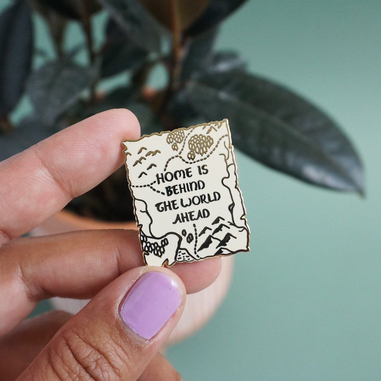 A small gold enamel pin of a map in the style of the lord of the rings with the words "Home is behind, the world ahead" on it.