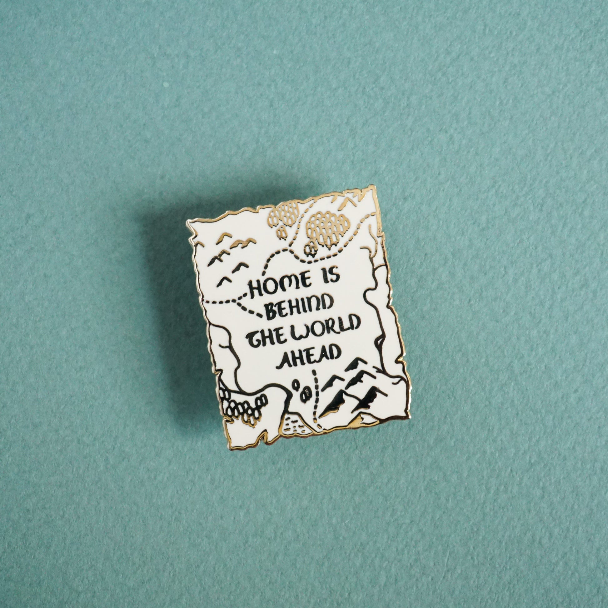 Small white enamel pin with gold metal and text that reads home is behind the world ahead