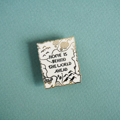 Small white enamel pin with gold metal and text that reads home is behind the world ahead