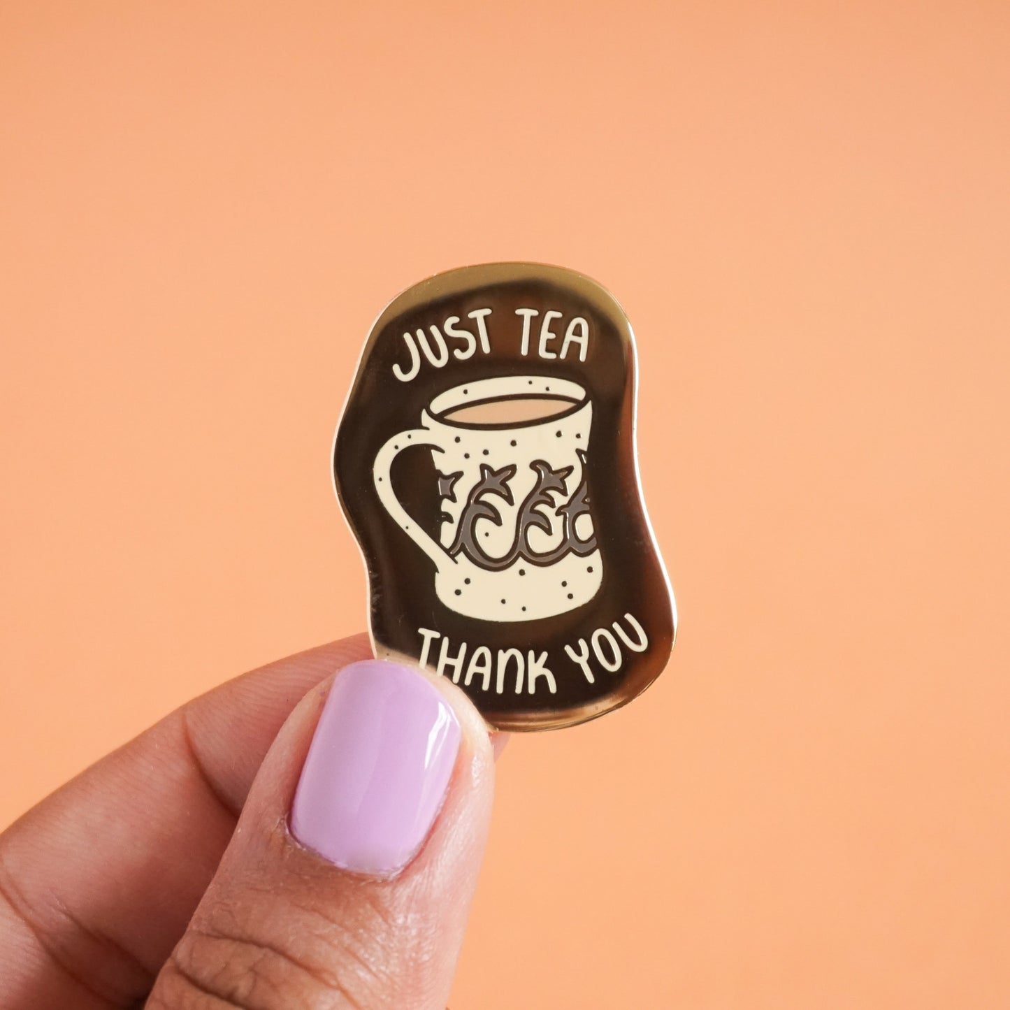 A small gold enamel pin with the words "just tea thank you" on it around a vintage style mug with tea in it.