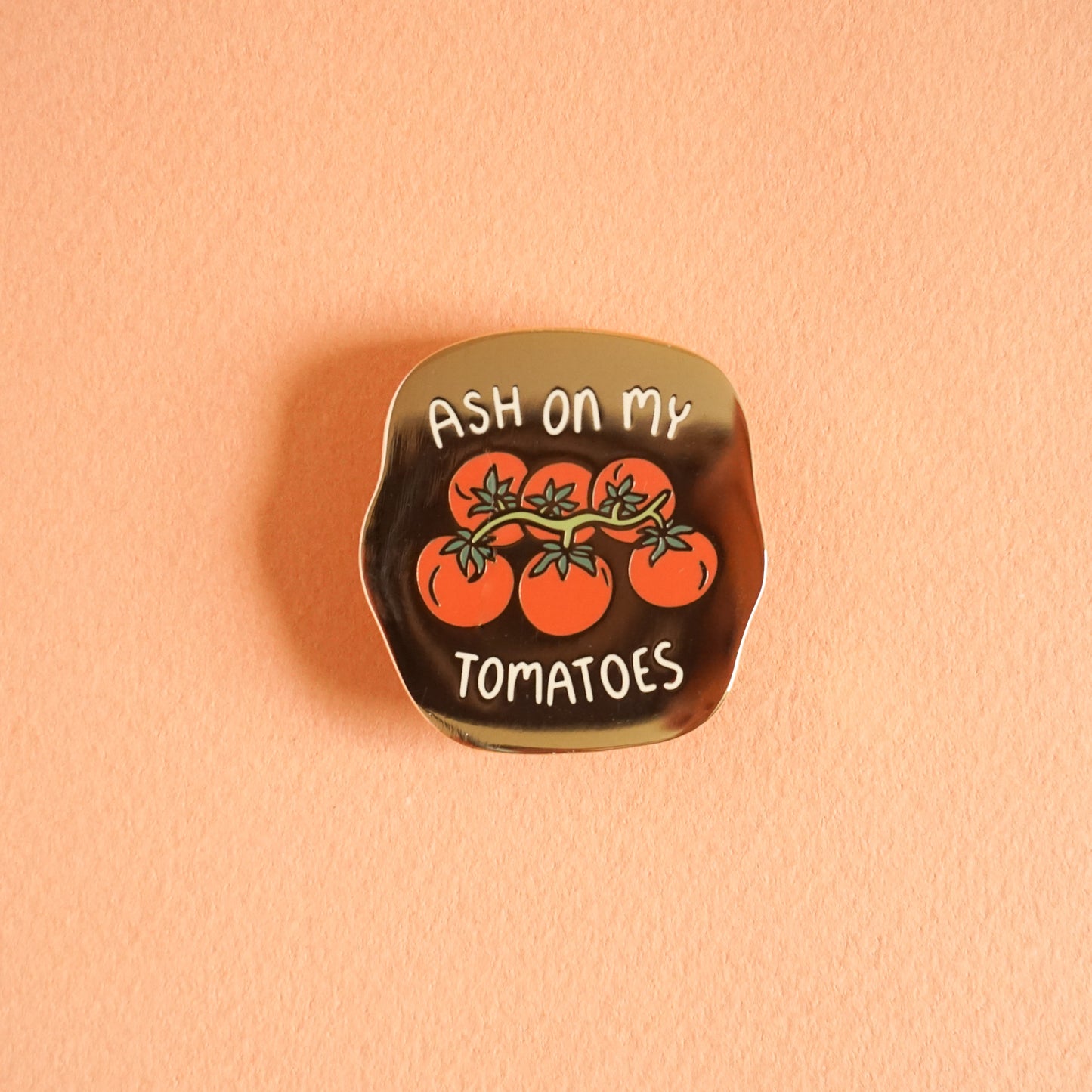 A small gold enamel pin that says "ash on my tomatoes" with a bunch of grape tomatoes on the vine in the center.