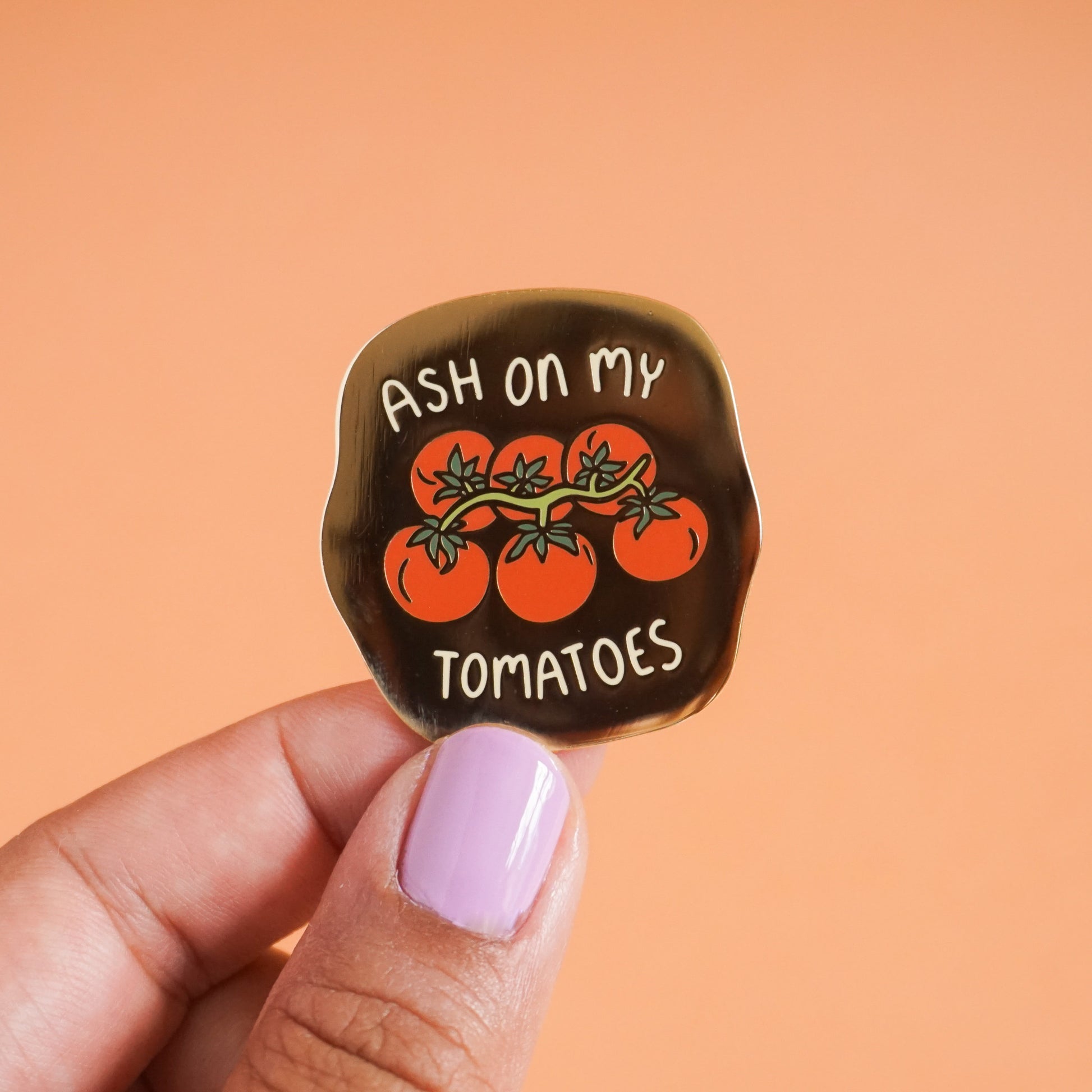 A small gold enamel pin that says "ash on my tomatoes" with a bunch of grape tomatoes on the vine in the center.