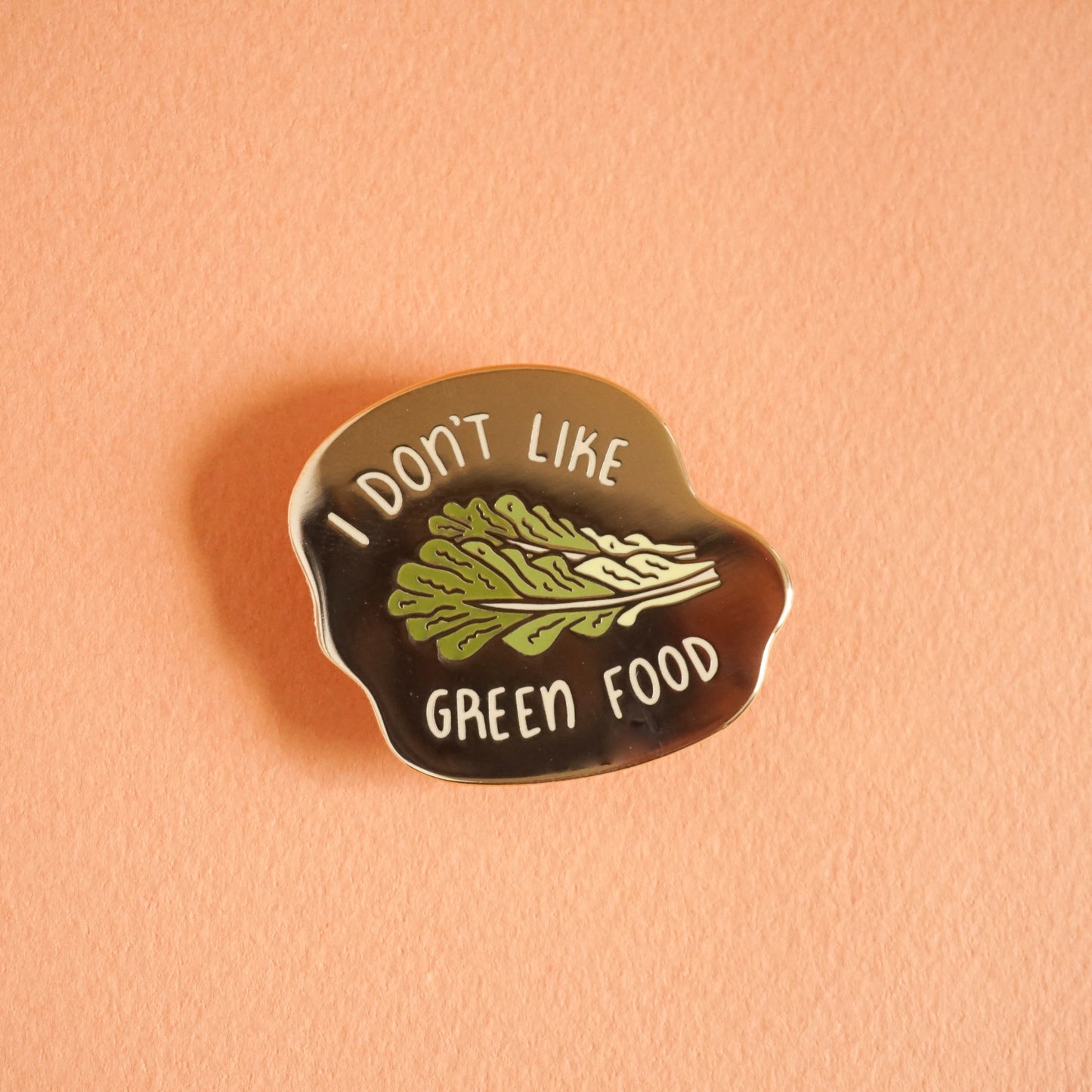 A small gold enamel pin that says "I don't like green food" with a romaine lettuce leaf in the center.