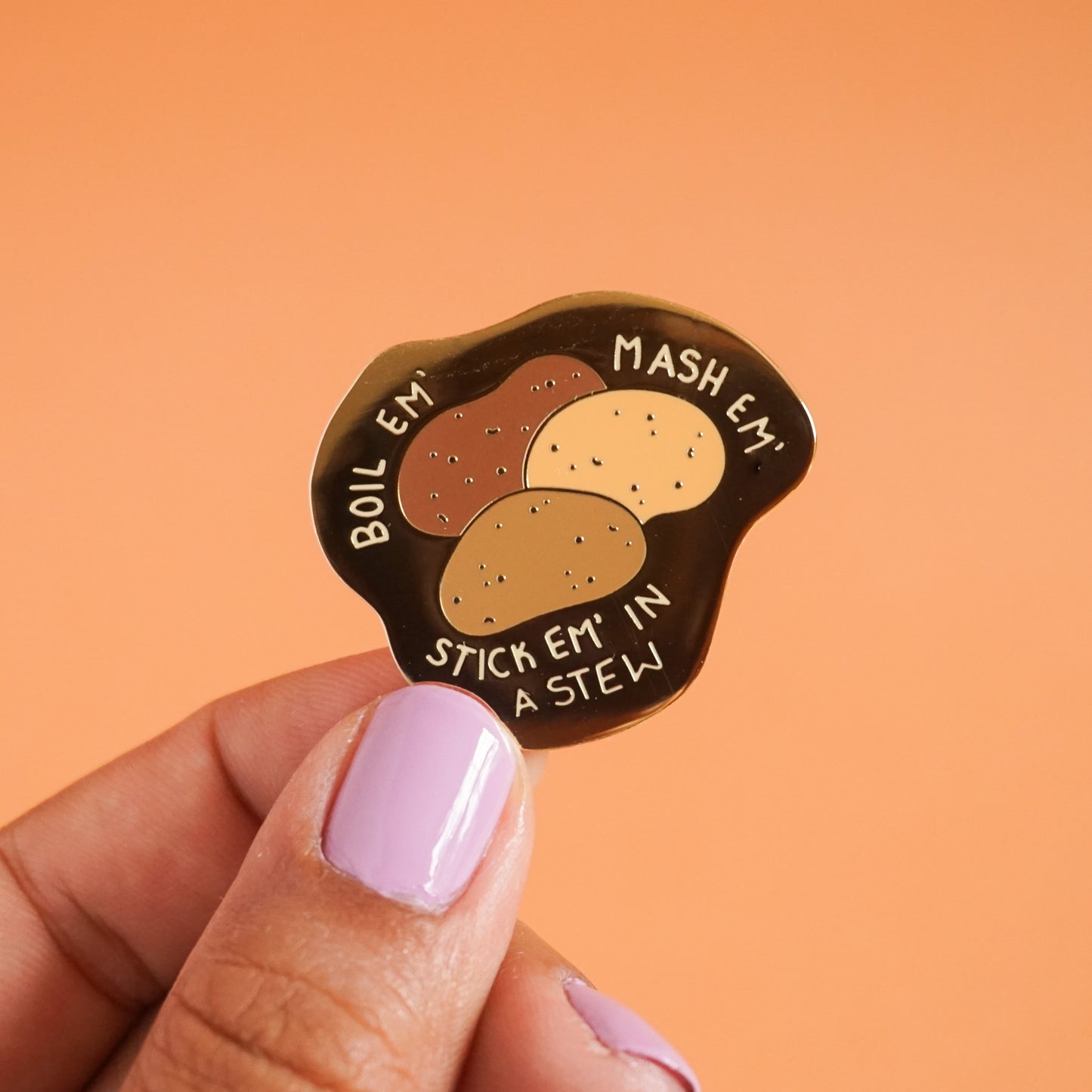 A small enamel pin that says "boil em, mash em, stick em in a stew," around some different colored potatoes.