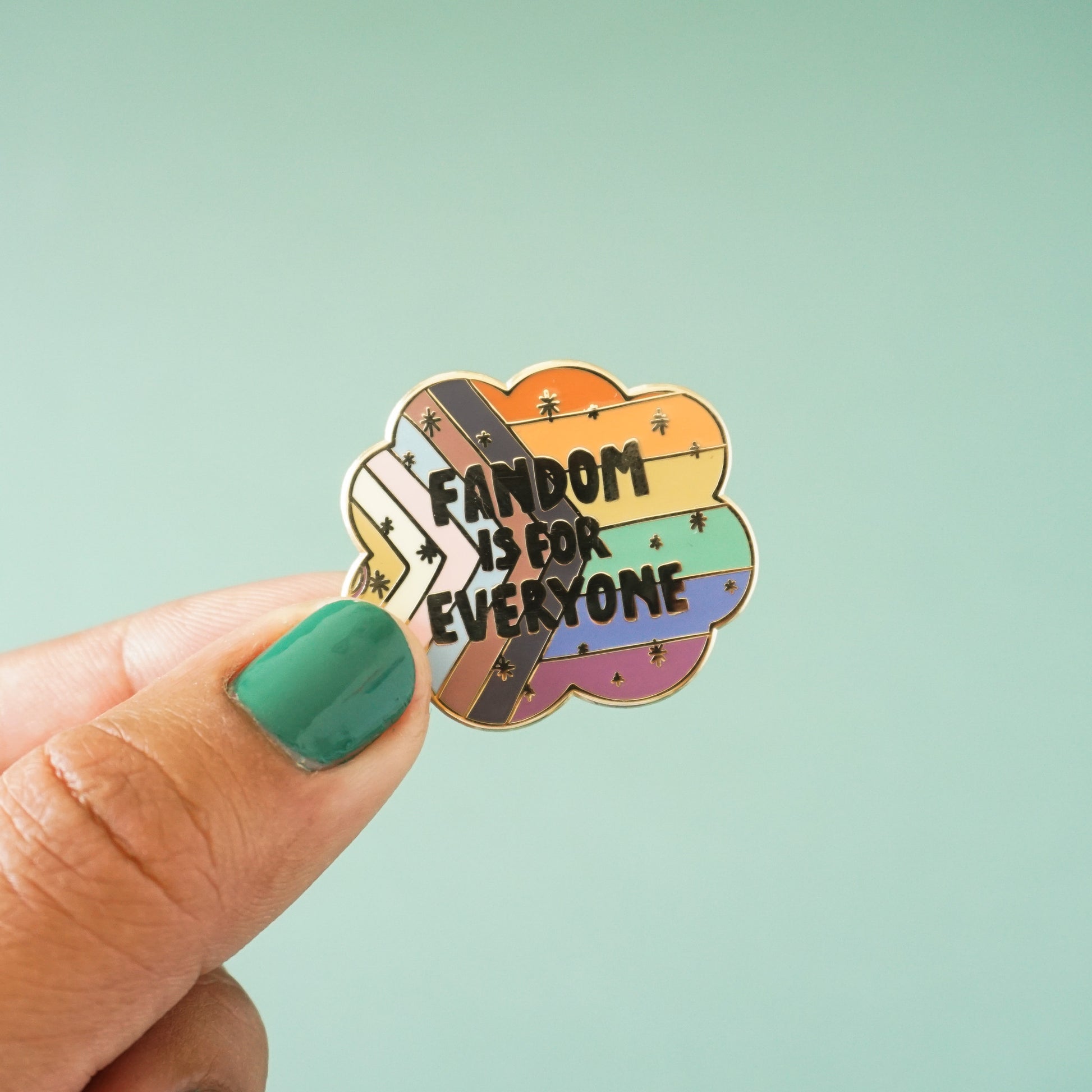 A gold enamel lapel pin showing the LGBTQIA+ pride flag with the words "Fandom is for everyone" over top.