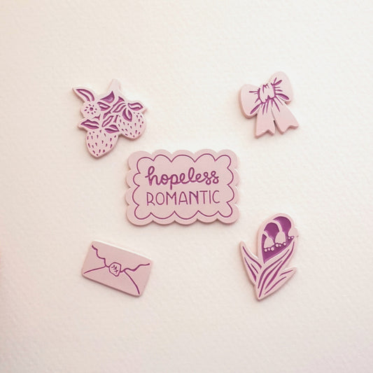 five light pink enamel pins of a bow, strawberries, lillies of the valley, an envelope, and the words "hopeless romantic"