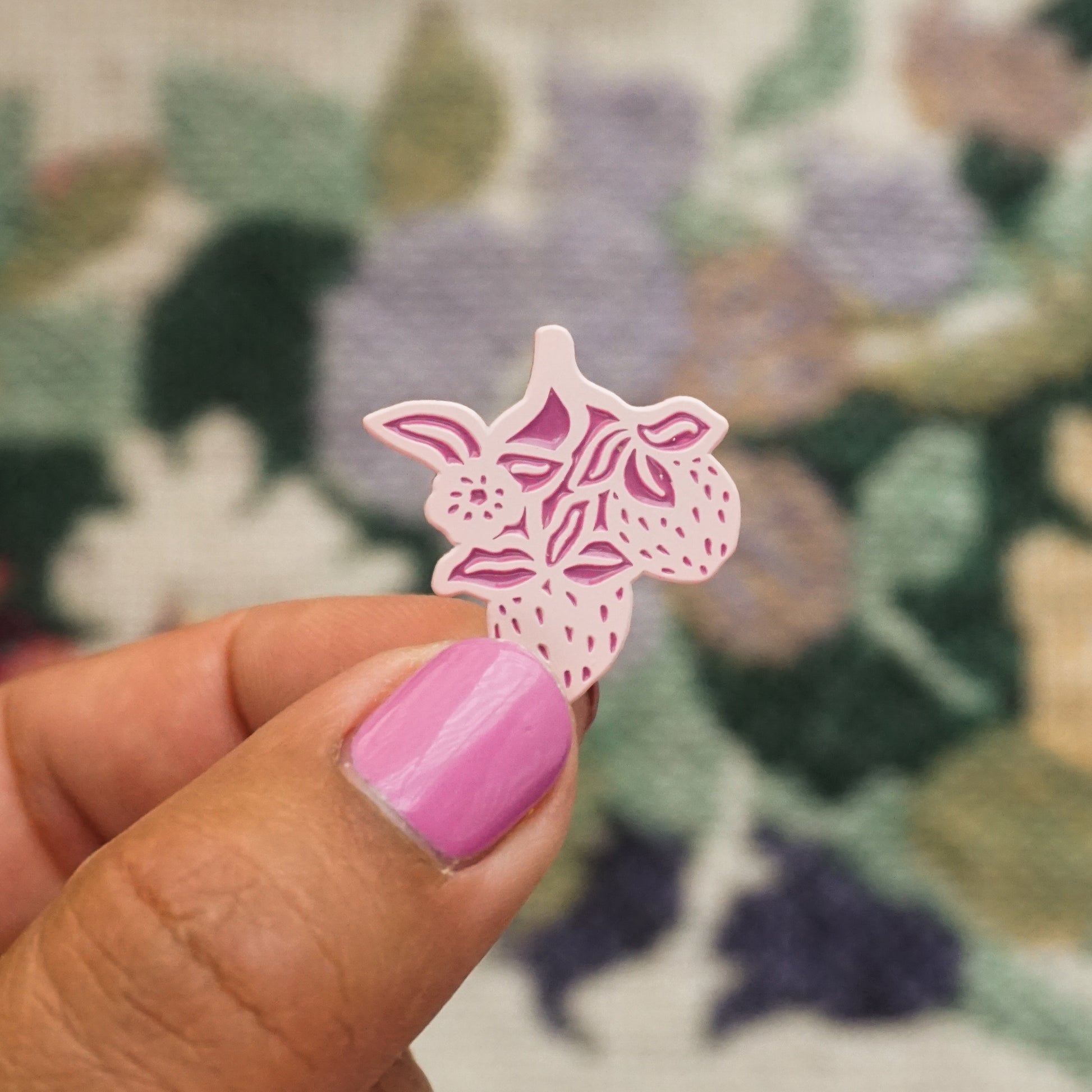 a hand holds up a light pink soft enamel pin in the shape of strawberries and a strawberry blossom
