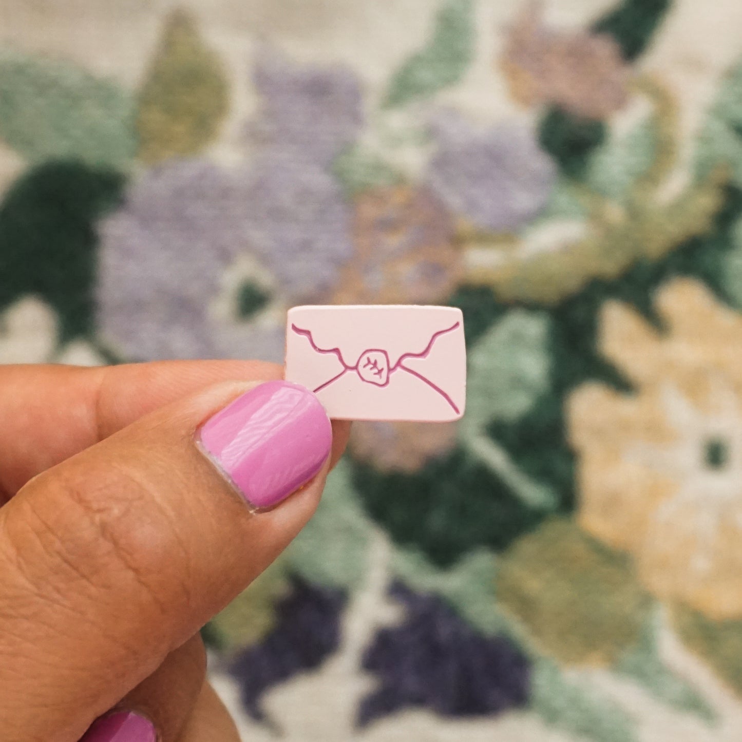 a hand holds up a light pink soft enamel pin in the an envelope with a scallop edged flap