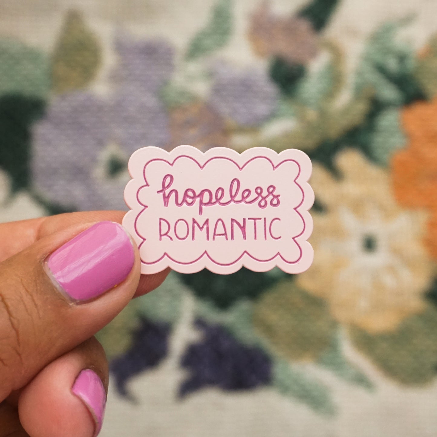 a hand holds up a light pink soft enamel pin with scalloped edges and the words "hopeless romantic"