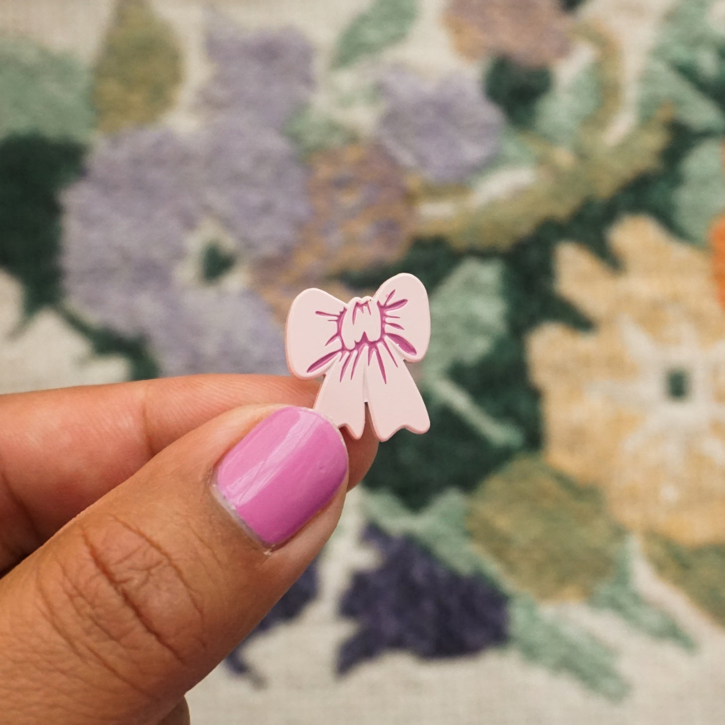a hand holds up a light pink soft enamel pin in the shape of a bow
