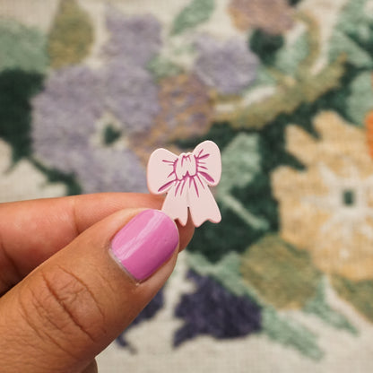 a hand holds up a light pink soft enamel pin in the shape of a bow