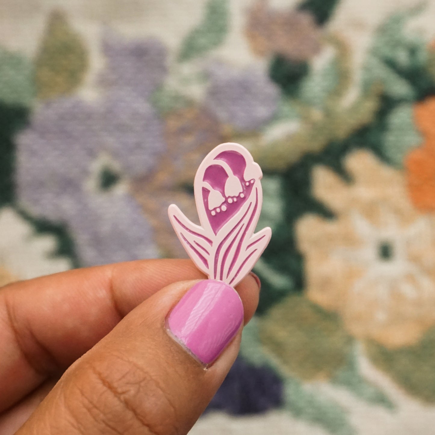 a hand holds up a light pink soft enamel pin in the shape of a lily of the valley plant with flowers