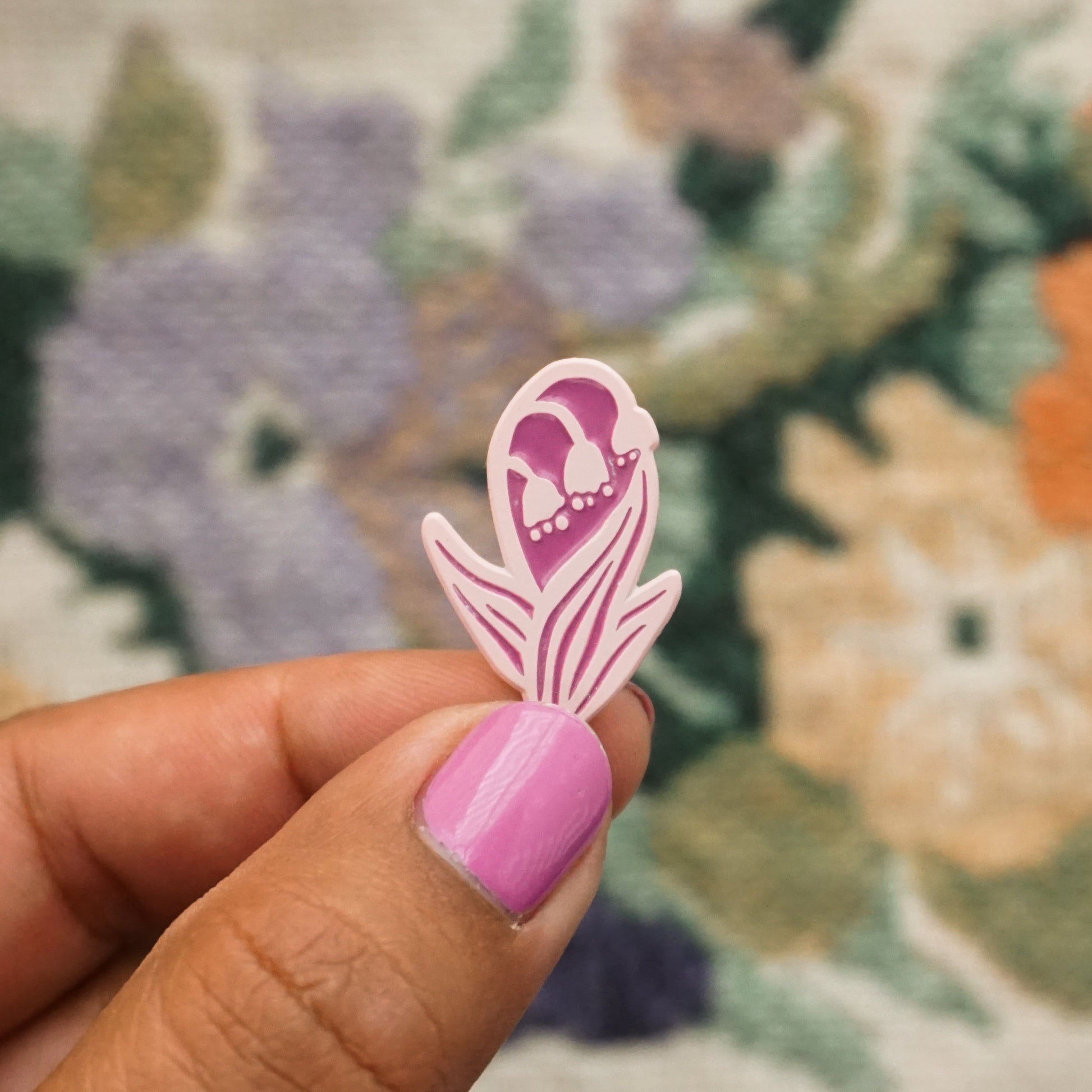 a hand holds up a light pink soft enamel pin in the shape of a lily of the valley plant with flowers