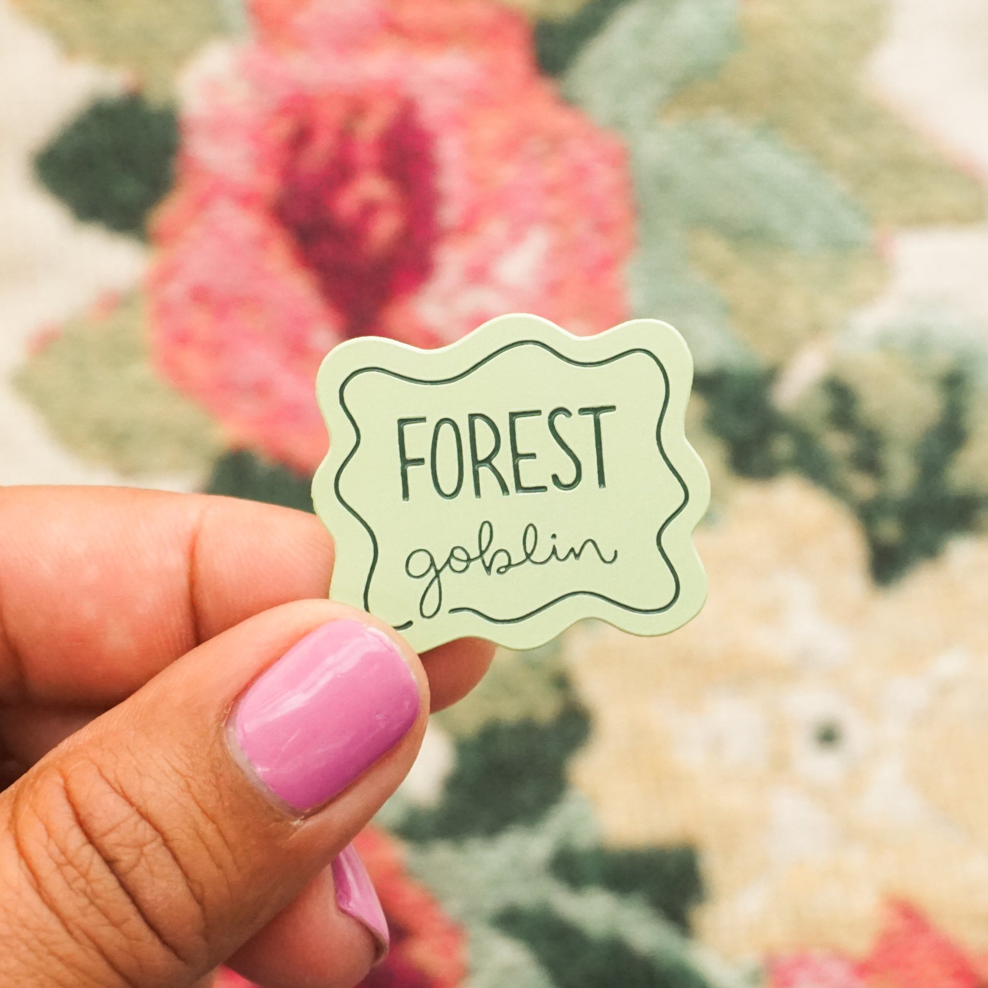a hand holds up a mint green pin that says "forest goblin" with wavy edges on the enamel pin