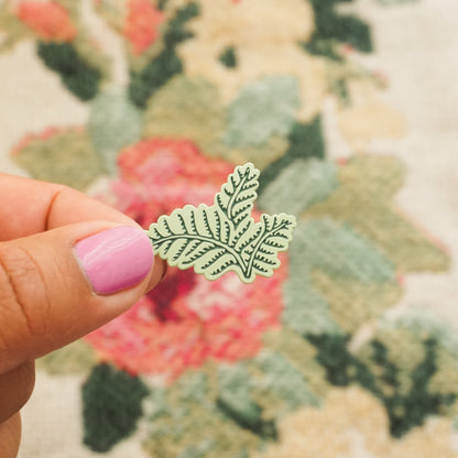 a hand holds up a mint green pin in the shape of a fern bush