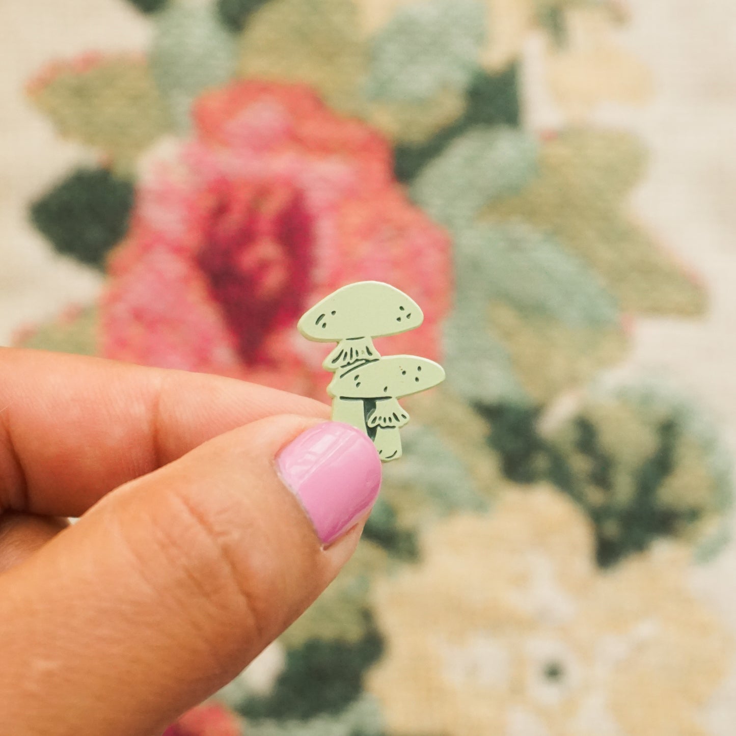 a hand holds up a mint green pin in the shape of two mushrooms