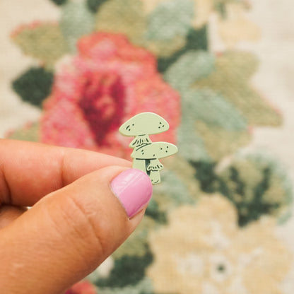 a hand holds up a mint green pin in the shape of two mushrooms