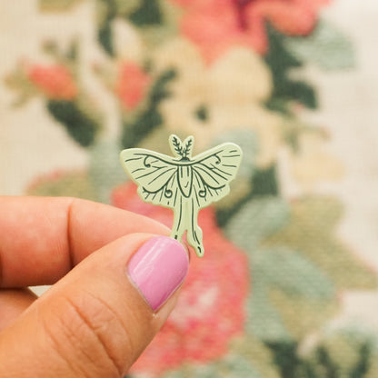 a hand holds up a mint green pin in the shape of a luna moh