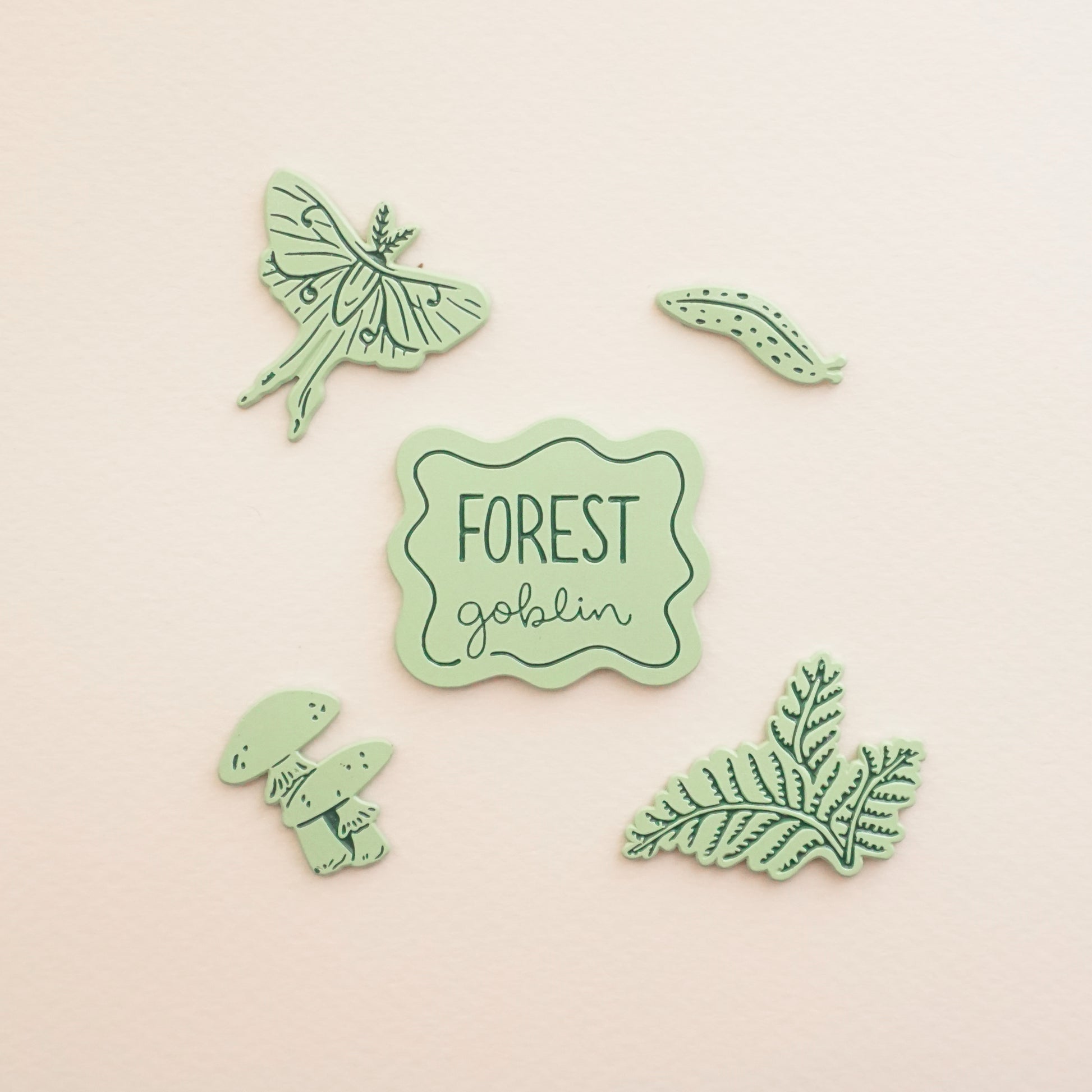 five light green pins of a slug, ferns, a luna moth, mushrooms, and a pin that says forest goblin