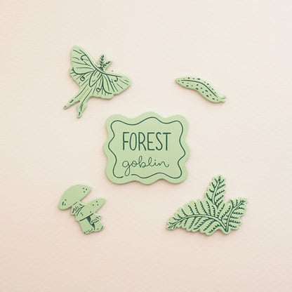 five light green pins of a slug, ferns, a luna moth, mushrooms, and a pin that says forest goblin