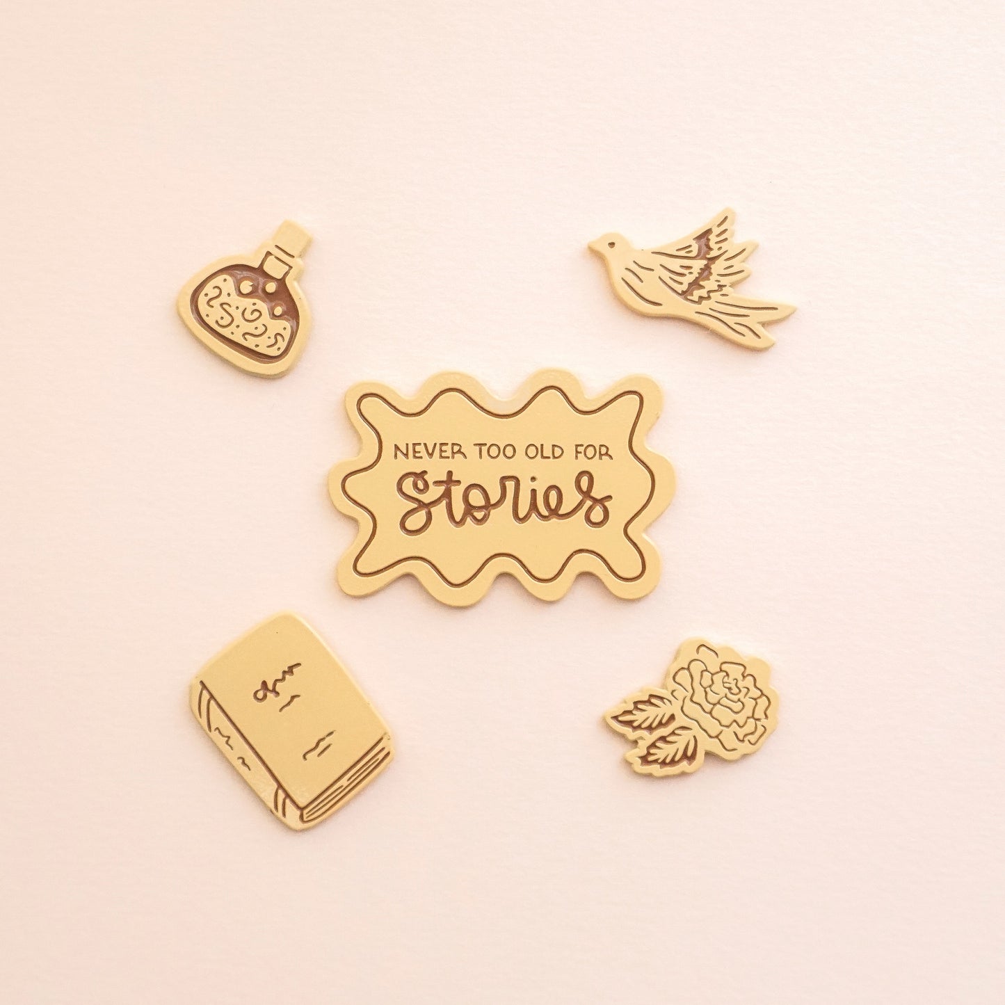 four peachy colored enamel pins in the shape of a potion bottle, pigeon, book, and rose, and the fifth pin says "never too old for stories