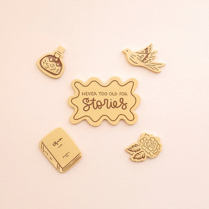 four peachy colored enamel pins in the shape of a potion bottle, pigeon, book, and rose, and the fifth pin says "never too old for stories