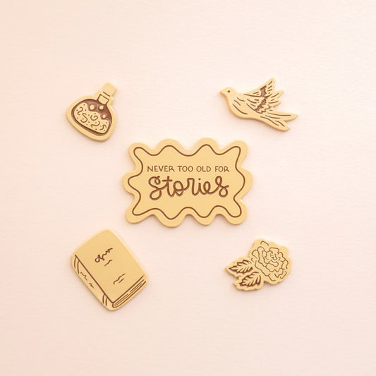 four peachy colored enamel pins in the shape of a potion bottle, pigeon, book, and rose, and the fifth pin says "never too old for stories