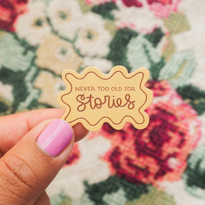 hand holding a peach colored enamel pin that says "never too old for stories"