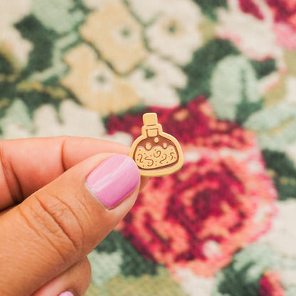hand holding a peach colored enamel pin in the shape of a potion bottle
