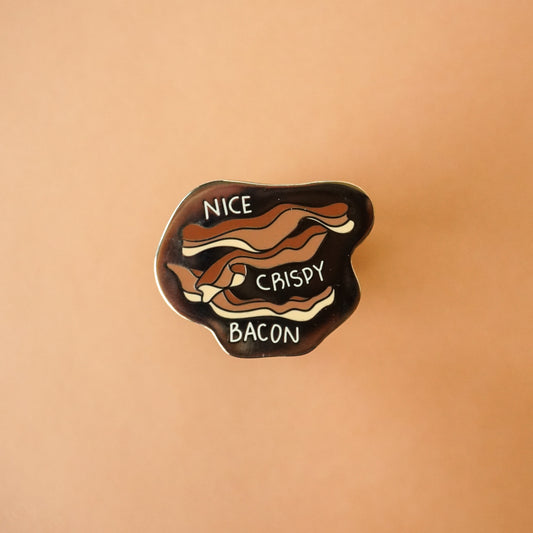 A small gold enamel pin with bacon on it and the words "nice, crispy, bacon."