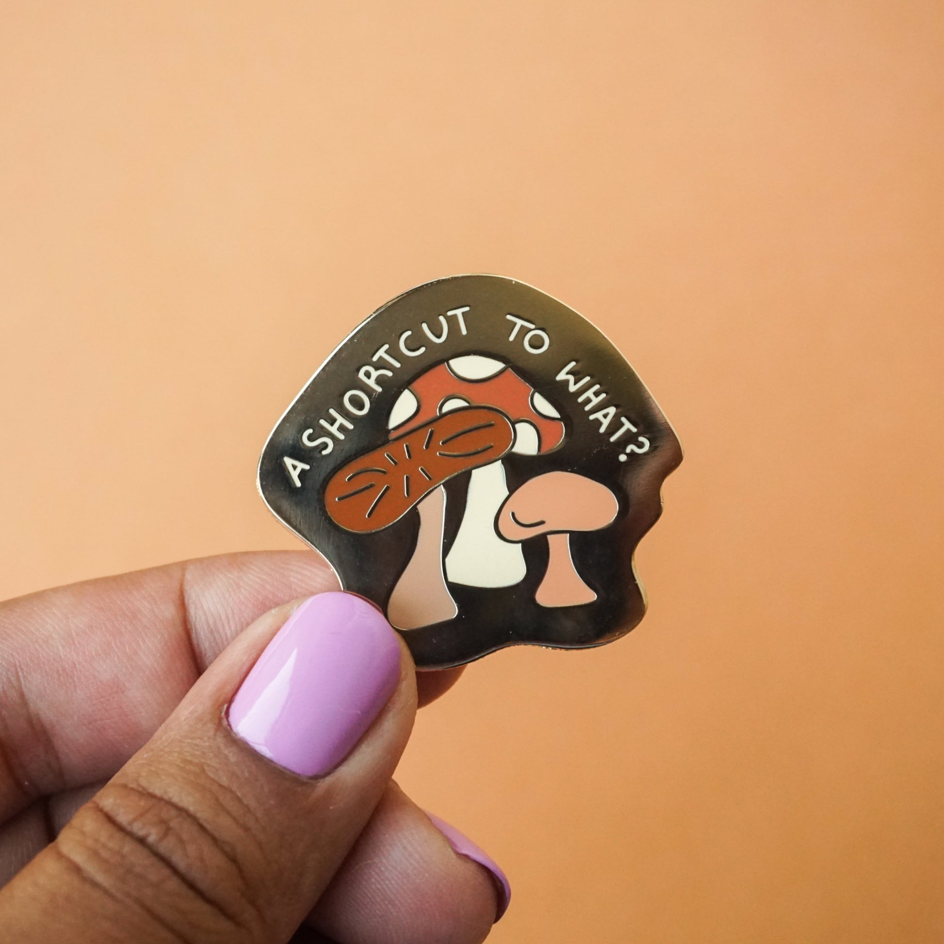 A gold enamel pin that says "a shortcut to what?" on it with mushrooms in it.