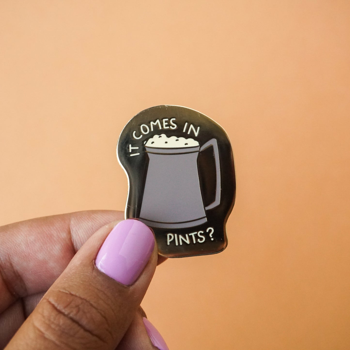 A small gold enamel pin with a pint of ale on it and the words "it comes in pints?"