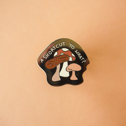 A gold enamel pin that says "a shortcut to what?" on it with mushrooms in it.