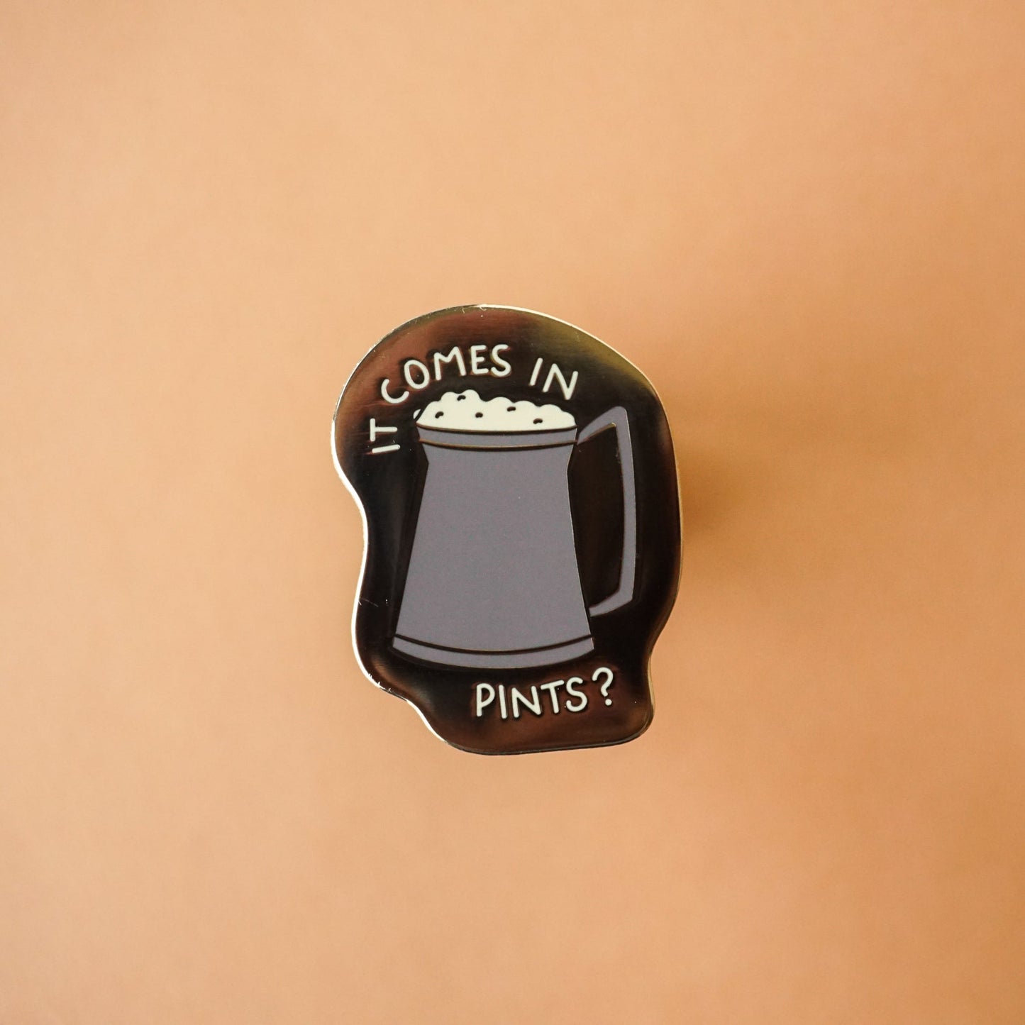 A small gold enamel pin with a pint of ale on it and the words "it comes in pints?"