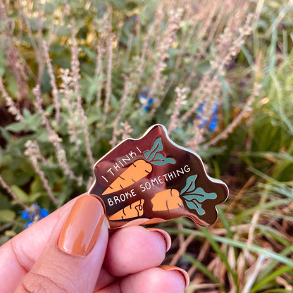 A hand holds up a gold enamel pin with the words "I think I broke something" with carots on the pin.