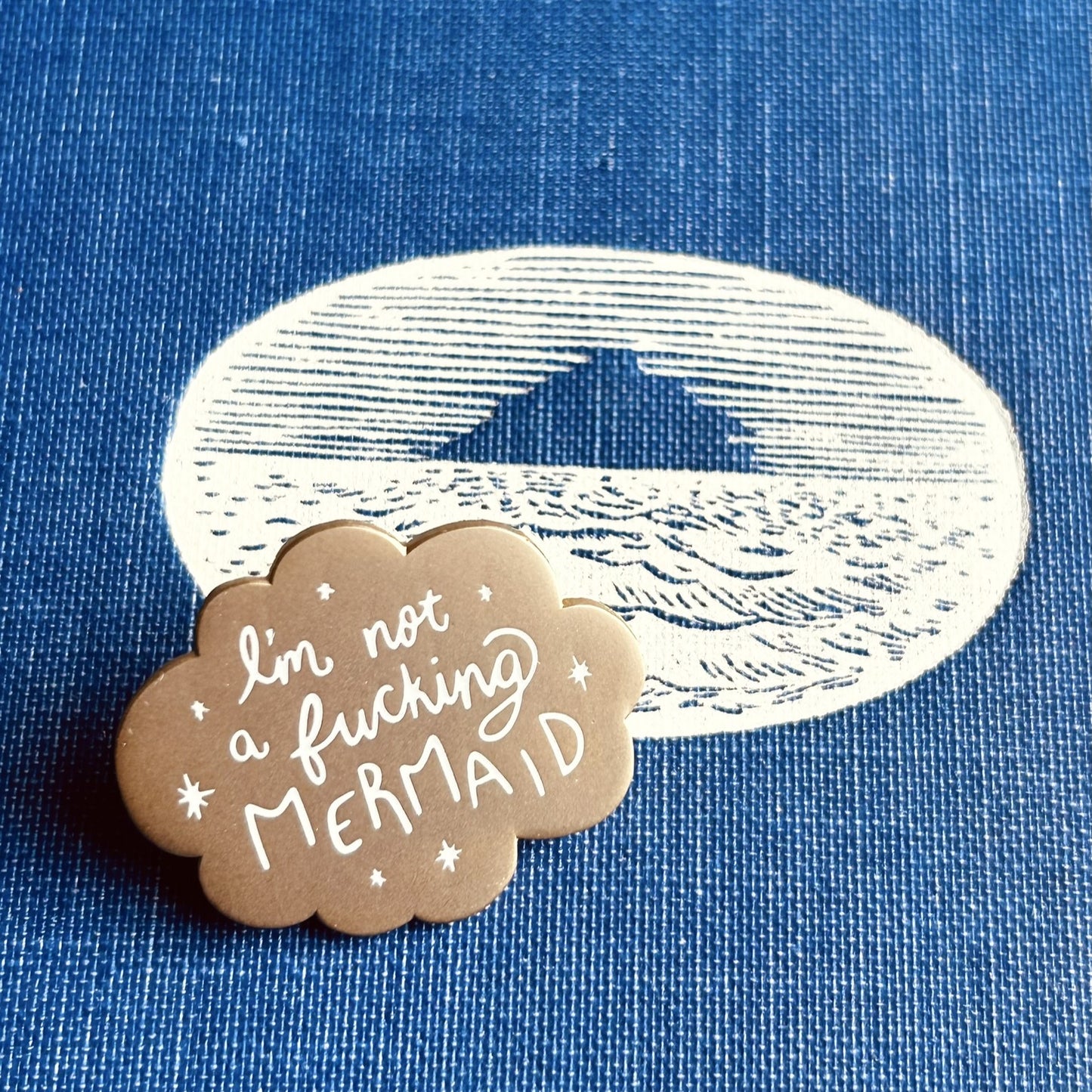 A small matte gold enamel pin with scalloped edges and white stars around the words "I'm not a fucking mermaid"