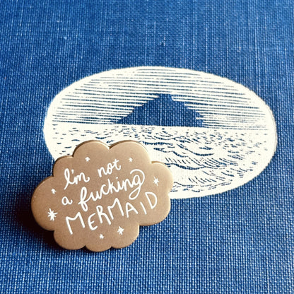 A small matte gold enamel pin with scalloped edges and white stars around the words "I'm not a fucking mermaid"