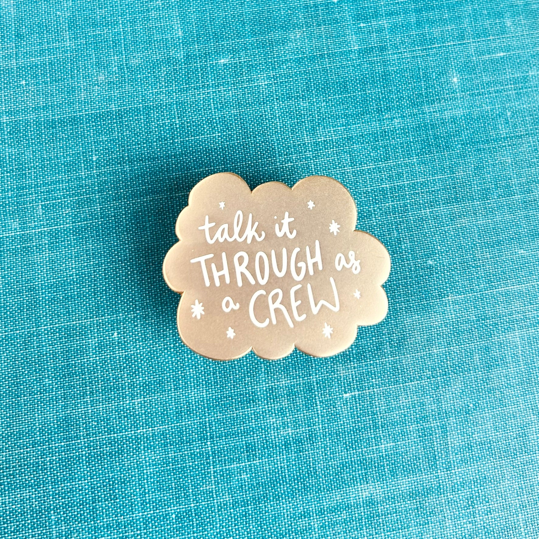 A small matte gold enamel pin with scalloped edges and white stars around the words "talk it through as a crew"